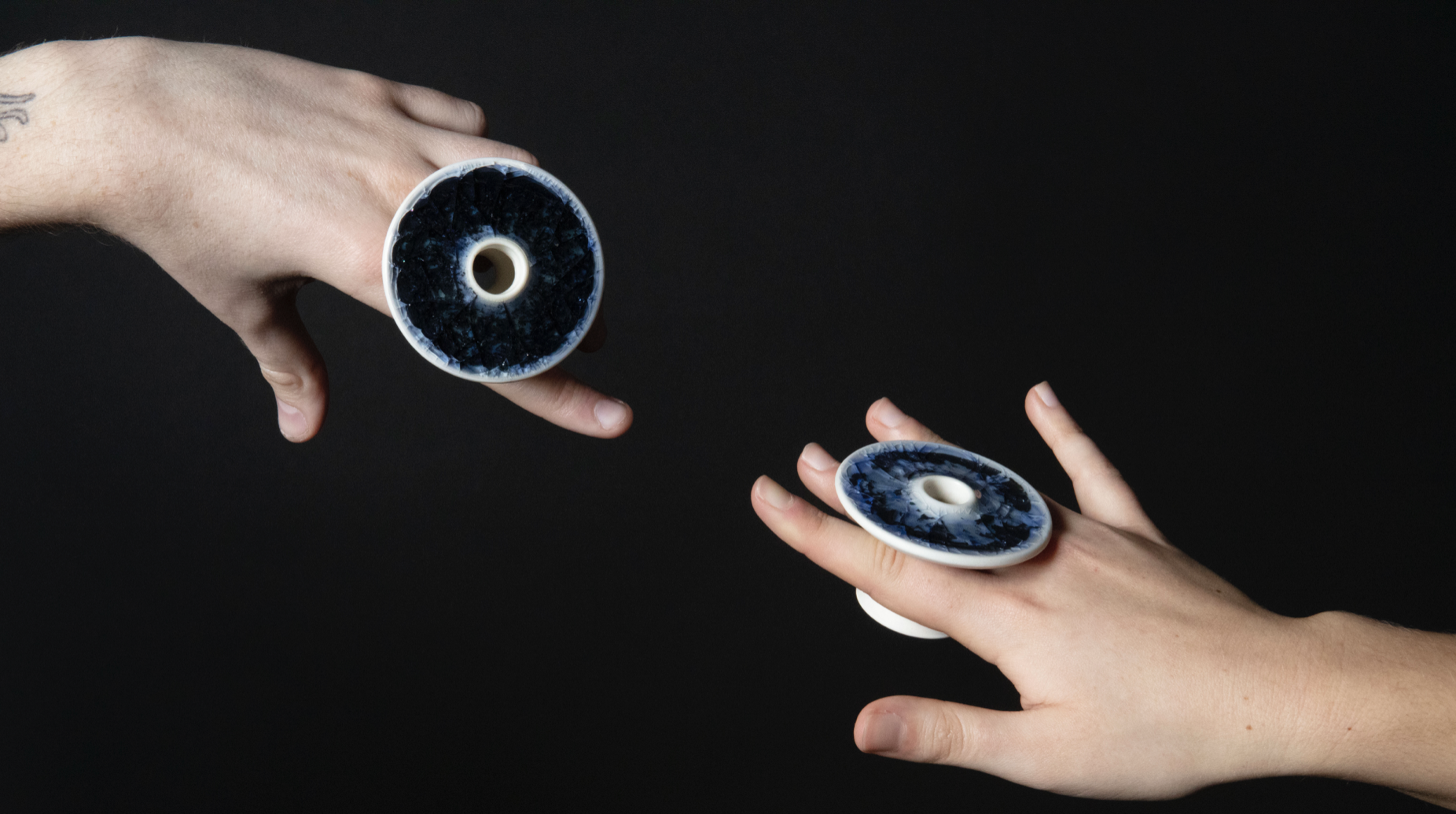 Two hand reach out from opposite ends of the image towards each other. The hands are against a deep black background and are holding a statement porcelain ring between the index and third finger. Rings by Zachary Dunlap, image by Katie Davies.