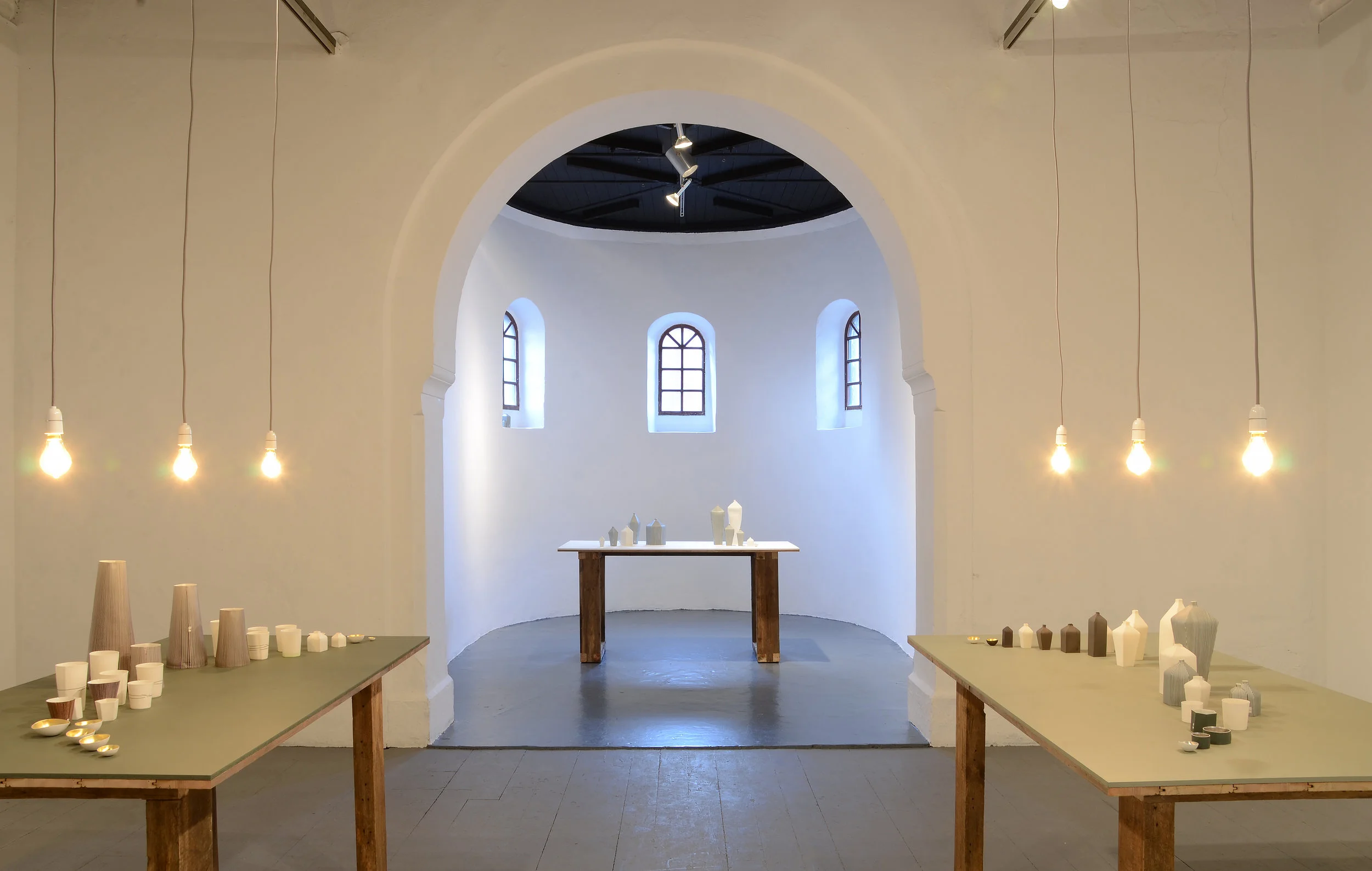 Delwedd o arddangosfa LLinellau Newidiol Justine Allison; rhan o raglen Iaith Clai. Mae yna dri bwrdd i'w gweld, dau yn y blaen ac un o fewn yr apse. Mae pob bwrdd â coesau pren trwchus, a'r top wedi eu paentio mewn gwhanol tonau o wyrdd. Mae gwaith serameg Justine ar bob bwrdd ac mae'r rhain wedi eu goleuo gan dri bwlb yn hongian uwchben bob bwrdd.